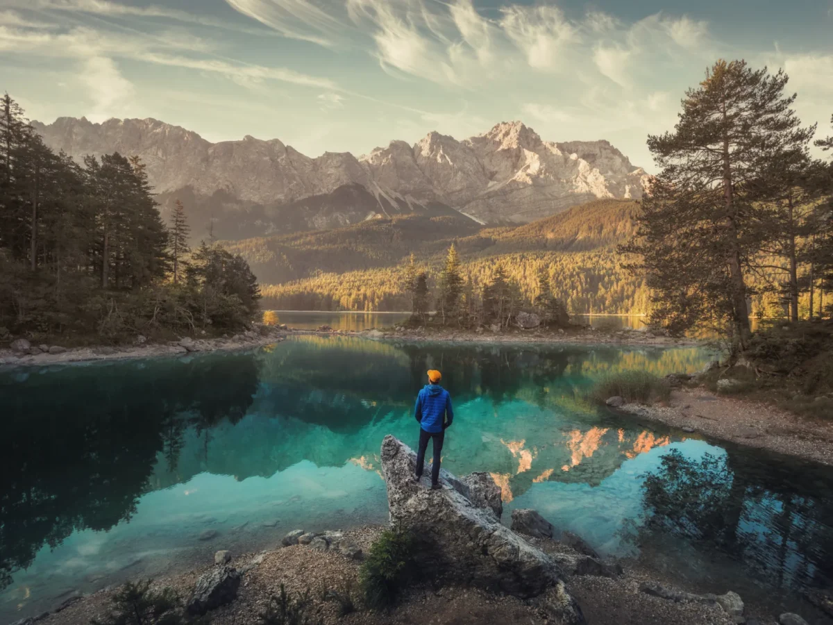 Person standing in front of a lake with mountain background reflecting in the water.