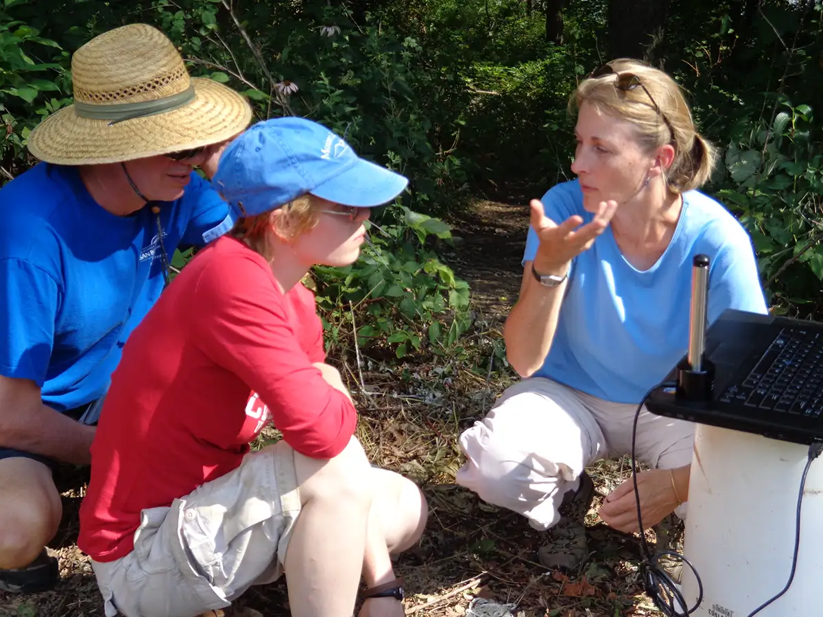Sue Swanson and two students in the field, crouched down in front of a laptop computer.
