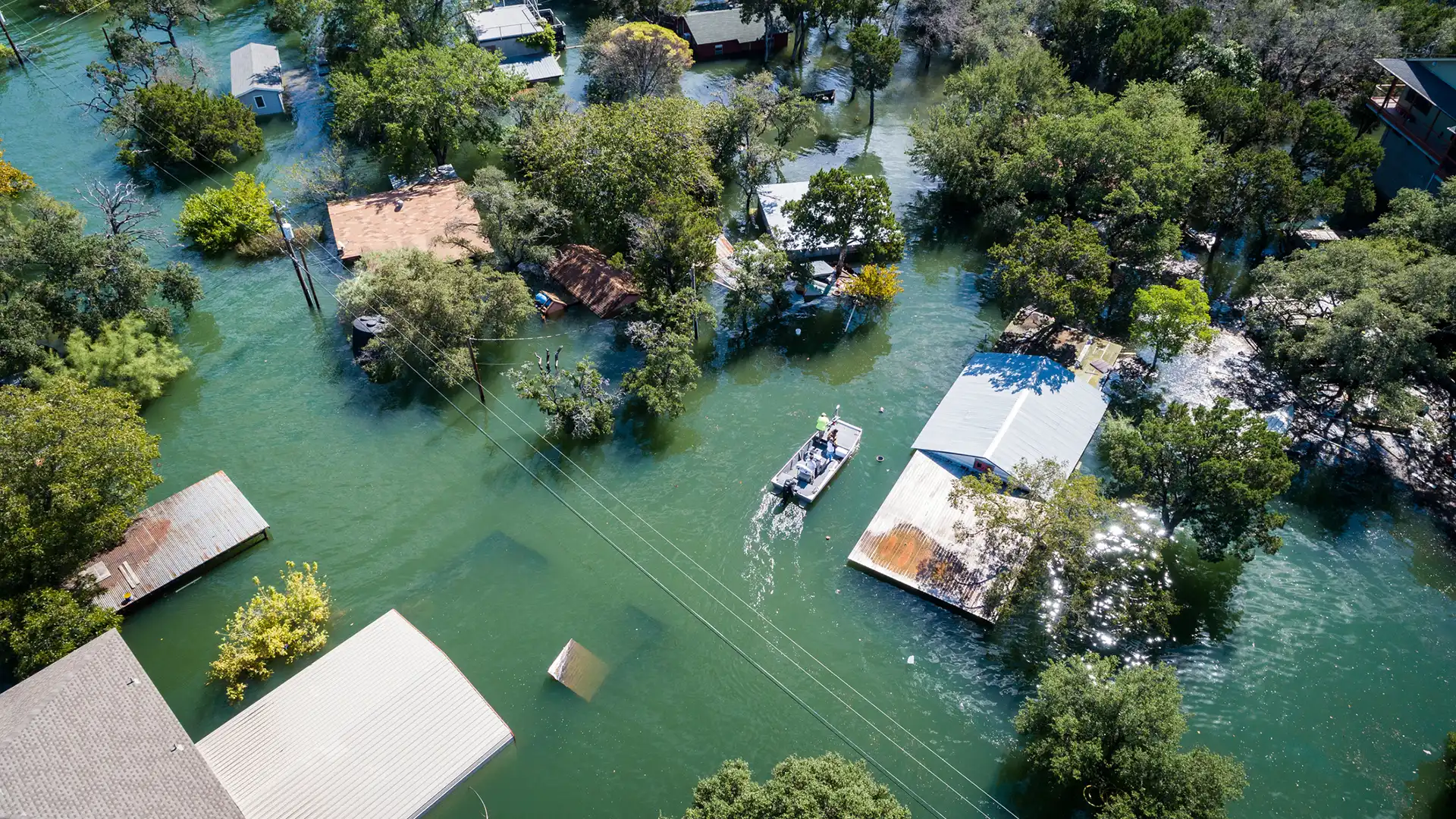 Flooded Texas neighborhood with homes submerged in water, trees partially underwater, and a rescue boat moving through the streets.