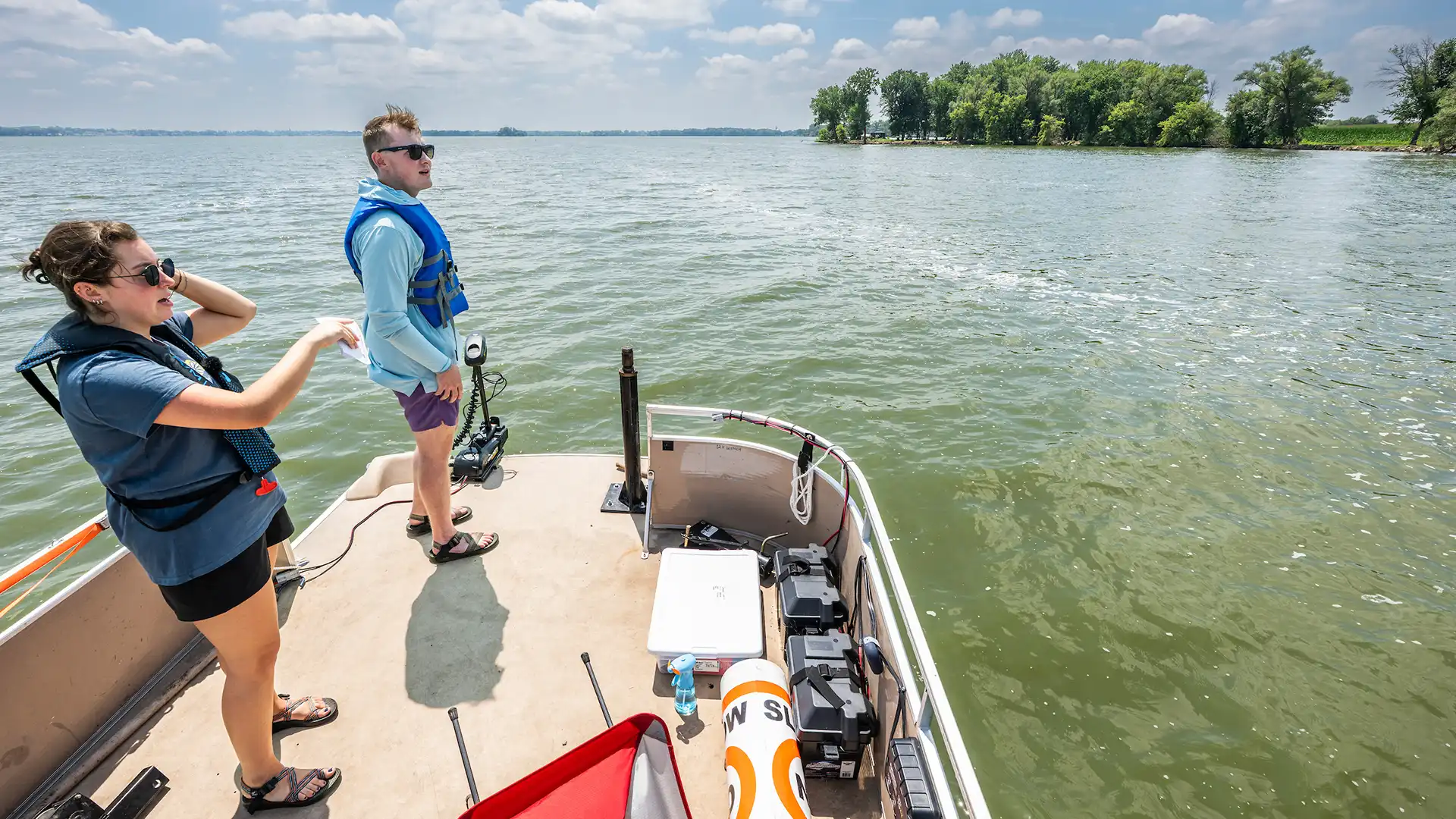 Two people standing on the end of a pontoon boat looking at a lake.