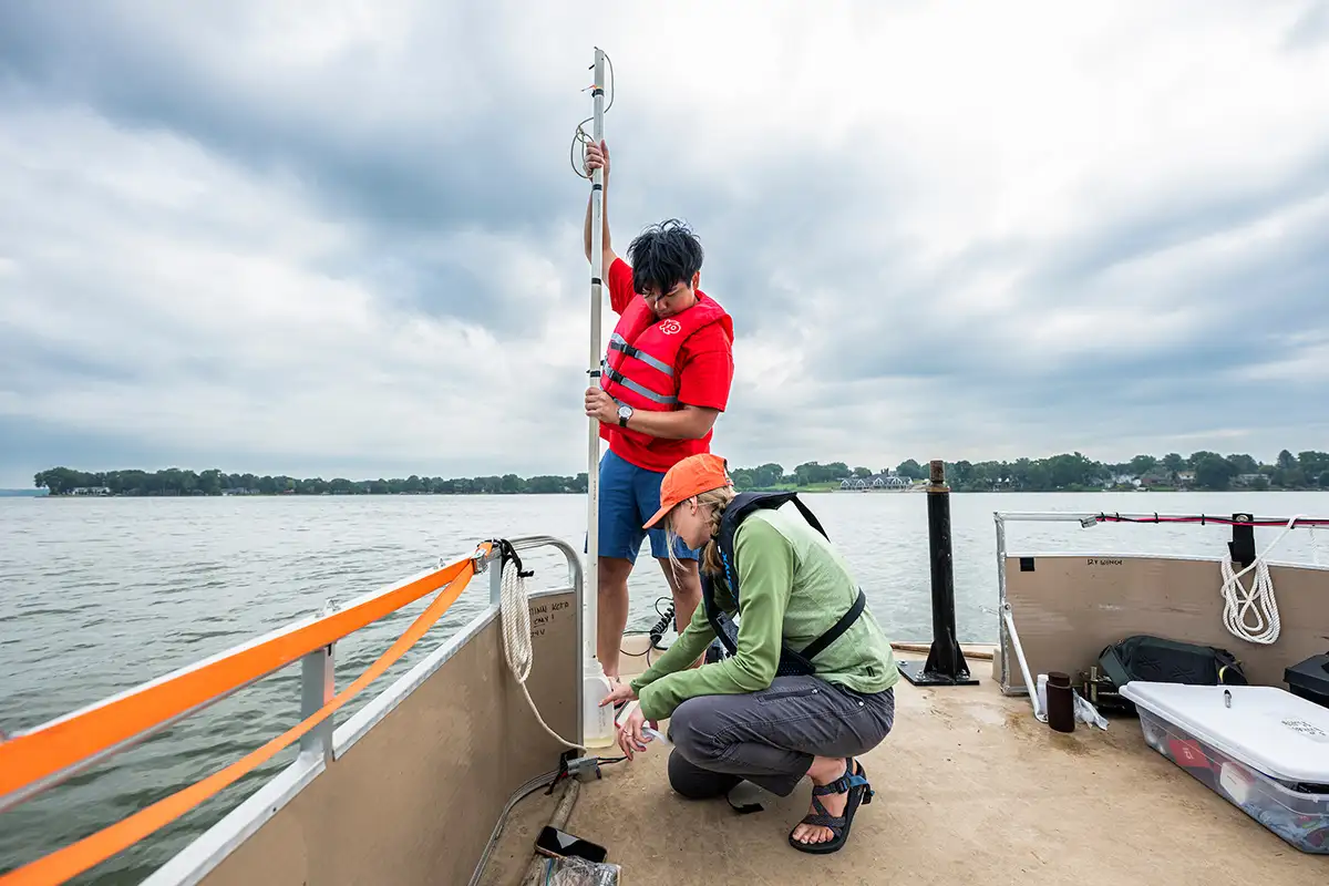 Two students standing on the end of a pontoon boat on a calm lake, holding a long rod used to collect water samples.