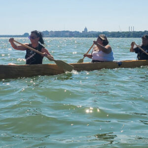 Three Ho-Chunk paddlers guide a dugout canoe, flying the Ho-Chunk flag, across Teewąąšikhomįik (Lake Mendota). Photos courtesy of Jessie Conaway