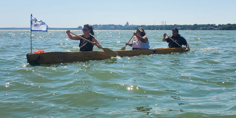 Three Ho-Chunk paddlers guide a dugout canoe, flying the Ho-Chunk flag, across Teewąąšikhomįik (Lake Mendota). Photos courtesy of Jessie Conaway