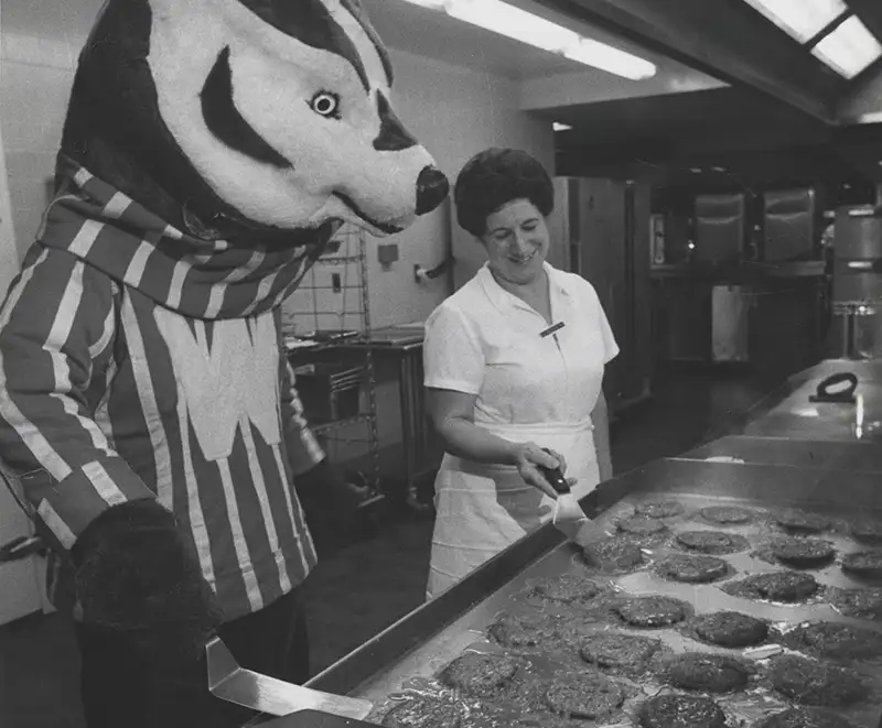 A cook teaches Bucky Badger how to cook hamburgers in a campus kitchen.