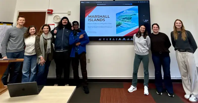 Group photo of students and instructors standing around a wall-mounted TV displaying a PowerPoint slide that says "Marshall Islands."