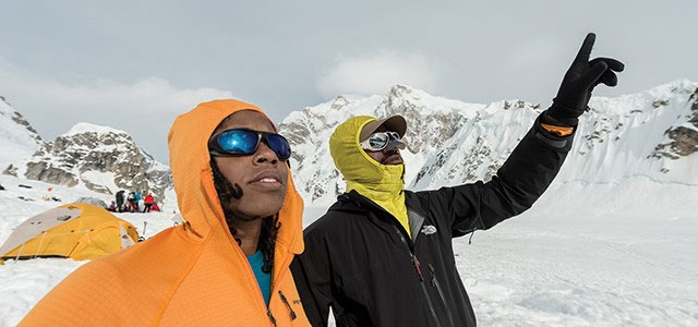 Two people standing amid a snowy mountainous landscape, dressed in winter gear and sunglasses and looking up at a bright sky