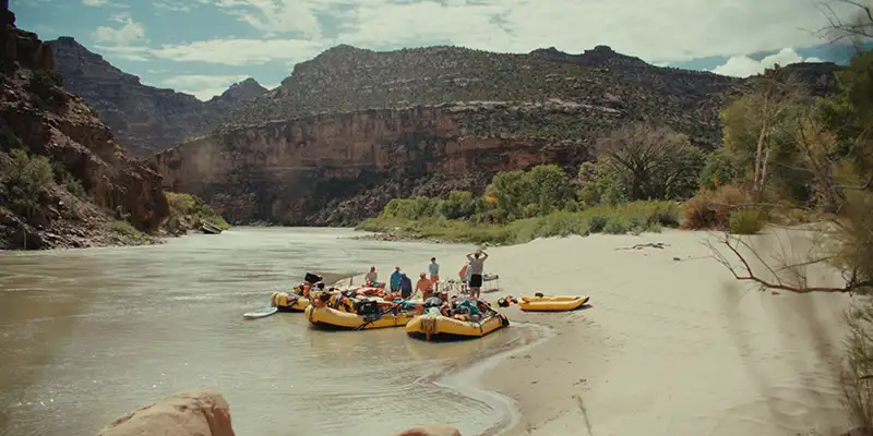 A group of people and their inflatable rafts gather on the sandy shore of a river with a majestic canyon in the background.