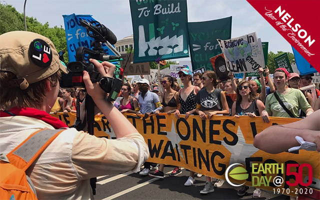 A large crowd of young people marching behind a banner that spans the width of a street